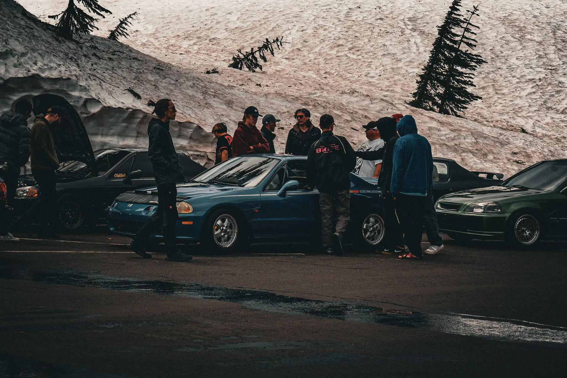 A group of people, dressed in winter clothing, stands around a blue car parked on a snow-covered mountain road during the Pacific Northwest Meet 2024. Other cars are parked nearby, with pine trees and snow-covered slopes creating a picturesque backdrop.