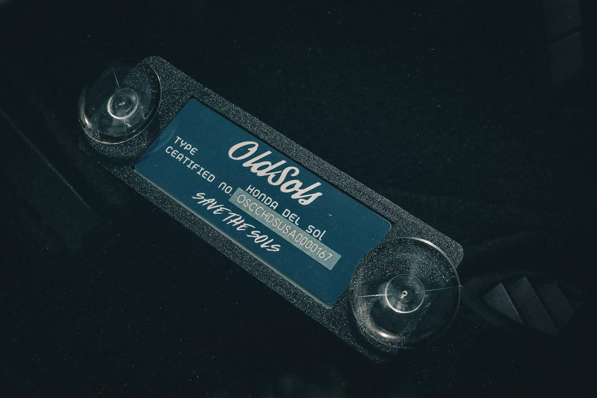 Close-up of a rectangular metal plate affixed to a surface with two clear suction cups. The plate has text in cursive and print that reads "OldSols," "TYPE HONDA CR-X DEL SOL," followed by a serial number and "Certified No. 00SCHDSOLS00000167" with "Pacific Northwest Meet 2024 - SAVE THE SOLS" at the bottom.