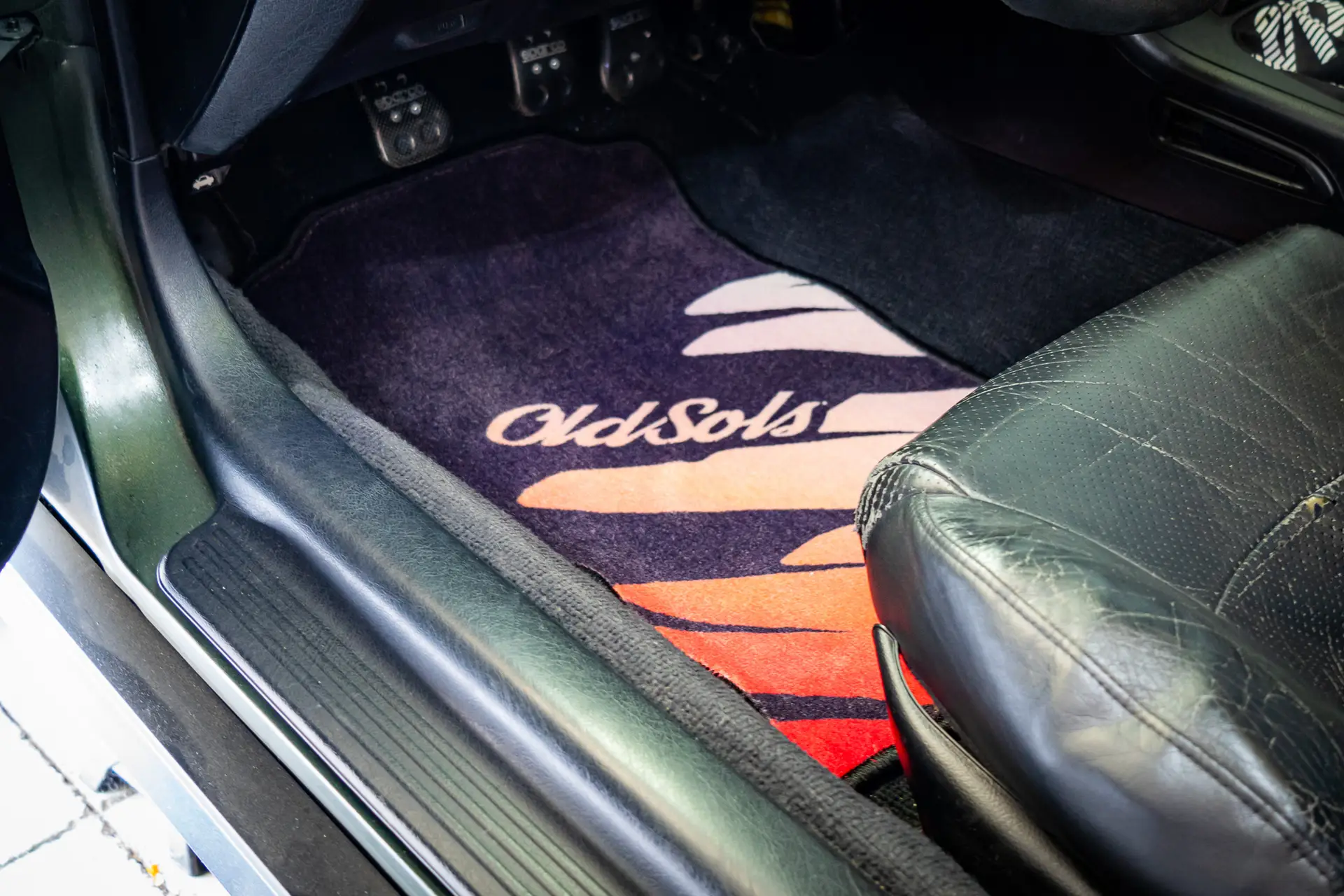 A vehicle's interior showcasing an OldSols Del Sol Floor Mat (LHD) featuring the text "OldSols" over a design of orange, red, and yellow sun rays. The black leather seat is partially visible, and the car's pedals can be seen in the background.